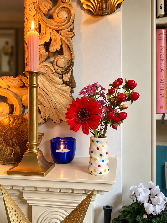 The Itsy-Bitsy Candle Holder holding flowers on a mantelpiece beside a brass candlestick and blue glass candle holder